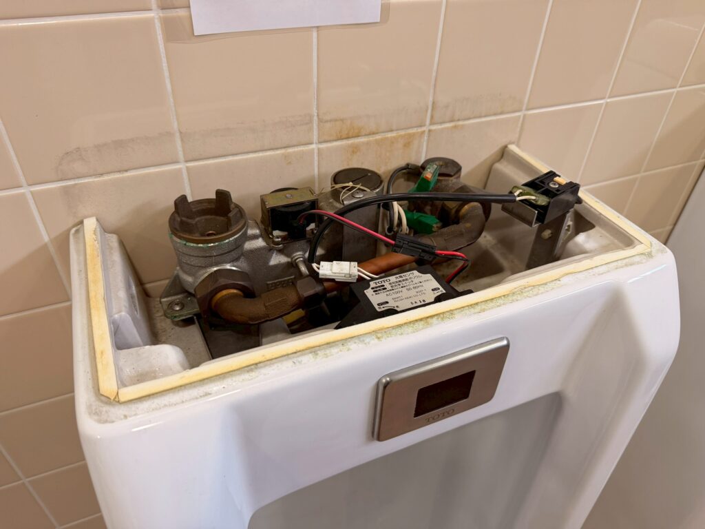 病棟の男子トイレの便器上部のカバーを取り外し、TOTO製のセンサーと電磁弁が見えている写真